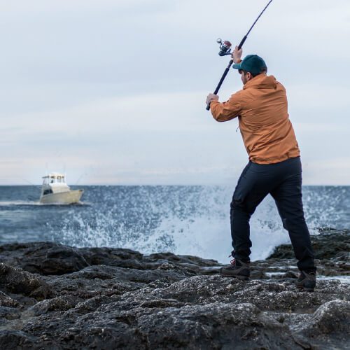 Man fishing on a rocky shore point with a boat in the background